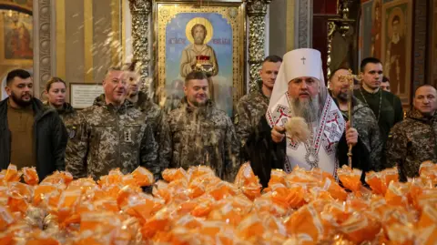 Metropolitan Epiphanius I, head of the Orthodox Church of Ukraine, sprinkles holy water on packets holding Easter cakes. Ukrainian service men stand behind him. 