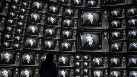 Real Ideas A person stands facing a curved wall filled with dozens of old-style television screens, each showing the same black-and-white image of a presenter speaking. The screens wrap around the space, creating an immersive display, with the viewer silhouetted against the glow of the repeated broadcasts.