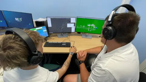 Mark Ansell/BBC Two other members of staff including Sam Johnson are sitting down in front of two computer screens. They are wearing headsets and controlling virtual planes for the simulator.
