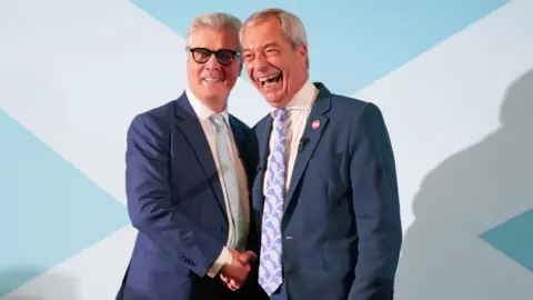 PA Media Malcolm Offord, who has grey hair and glasses, shakes hands with a smiling Nigel Farage, who has balding grey hair. Both are wearing dark suits, and standing in front of a screen with a white saltire cross on a pale blue background. 