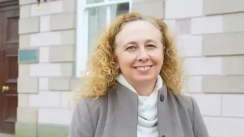 BBC Deputy Inna Gardiner smiles as she stands outside a stone building which has a large wooden door and a window. She has long and curly blonde hair and is wearing a grey jacket over a white jumper.