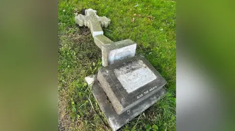 A gravestone shaped in a cross in a churchyard which has been smashed and fallen from the base on the grass.