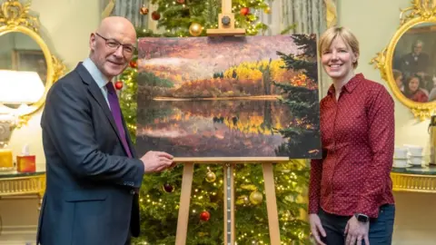 PA Media John Swinney standing in front of a Christmas tree with Jane Barlow and the winning photograph - an autumn scene of a loch with trees on a hillside.