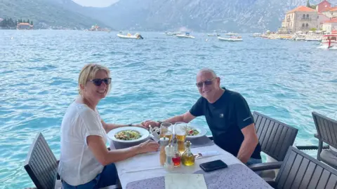 Jill Gale Jill and husband at a table in front of a wide blue lake, with villas on the distant shore and mountains behind the water. There are boats and dinghies dotted across the water.