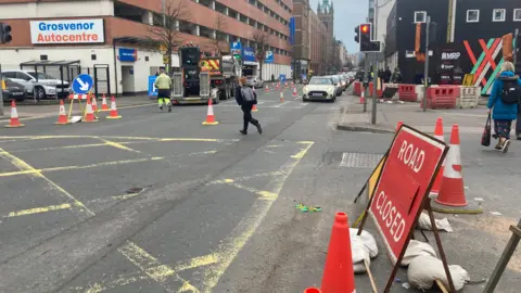 The junction of Grosvenor Road and Durham Street in Belfast.  There are traffic cones closing off one lane of Grosvenor Road and a "road closed" sign at the entrance to Durham street.  Pedestrians are crossing the roads and traffic is queuing at traffic lights. 