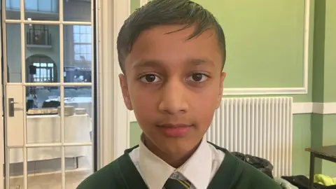 BBC Soham, a pupil at St Malachy's Primary school in Belfast. He has short, dark hair and is wearing a dark green school jumper, white shirt and a green and yellow striped tie.