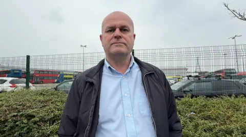 A bald man wearing a light blue shirt, and a dark blue jacket, stands in front of green fencing outside a depot.