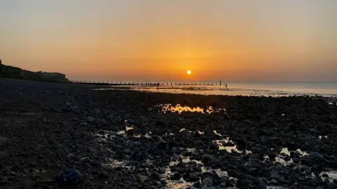 The sun dips down in to the sea off the coast of Cromer. Groynes in the sea are in silhouette and there are rocks in the foreground. The sky has hues of orange and grey.