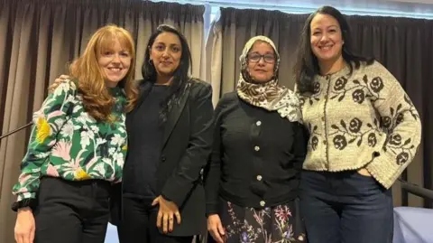 Four women stand closely together, smiling at the camera, after a panel discussion. Behind them are four chairs and banners for BBC Cymru Wales and Muslim Doctors Cymru.