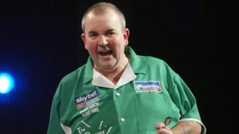 A man with short cropped hair is gripping a dart. He is wearing a green shirt with sponsor names on it
