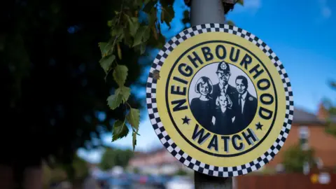 A round Neighbourhood Watch logo attached to a black lamp post. It is yellow and includes a black-and-white sketch of a police officer with three members of the community. The background, which is blurred, shows a leafy residential street.