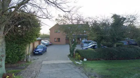 A modern two-storey brick house with brown window frames. There is a block-paved drive in front, where cars are parked, and garages to the left. There is a large tree to the left of the gateway and a hedge to the right.