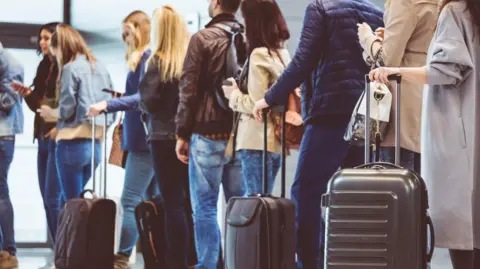 People queuing in airport with wheelie bags next to them