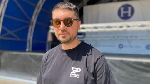 Alex Pope/BBC Gareth Barber, wearing a black T-shirt and sunglasses, standing by a stage. He is looking at the camera. 
