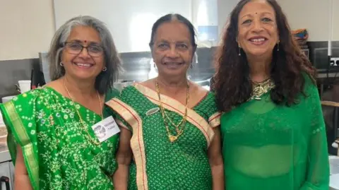 Three women are wearing green saris and are smiling in a kitchen. 