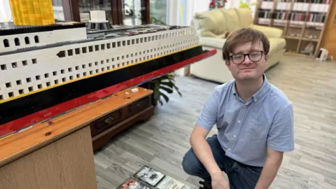 Jack Moore (a man with glasses and his hair in a curtain style, wearing a blue shirt) looks directly into the camera kneeling on his Lego creation of the titanic