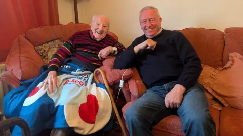 Alex Moss/BBC Two smiling men sat down next to each other. On the left is an older man, with balding light grey hair. He is wearing a striped coloured jumper and has a blanket over his knees with a walking stick resting to the side. On the right is a younger man, wearing dark blue jeans and a dark navy jumper. 