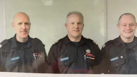 SFRS Three male firefighters standing in a row. They are all wearing black shirts with the Shropshire Fire and Rescue Service logo on the breast, with red writing. One is bald, one has short spiky grey hair and the other, Darren, has very short brown hair.