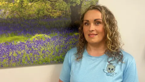 Sarah Owen has curly blonde hair, she wears a blue T-shirt which features a logo of an angel bear close to her left shoulder. She stands next to a large picture of bluebells.