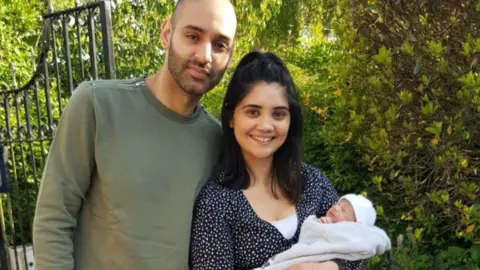RAJ CHAGGER Man in green jumper. Lady in black spotty top smiling holding a baby in blanket