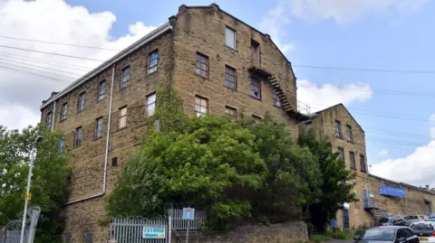 The former Victor Works woollen mills on Bolton Hall Road in Bradford which could be turned into flats. There are trees by the security gate and cars parked on the road.
