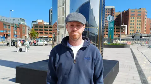 Kevin Killen is wearing a grey flat cap and a navy hoodie with a white top underneath. A large shiny sculpture is behind him. Some people in the background are pulling small suitcases. 