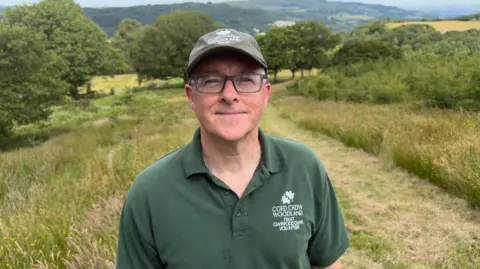 Keith stands smiling at the camera wearing a green top with the ancient woodland in behind him and the new tree saplings volunteers planted to the right of frame