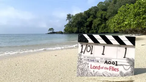 BBC/Eleven/Callum Devrell-Cameron A TV clapper board saying Lord of the Flies stuck in white sand on a beach with blue seas and a wooded cliff in the background