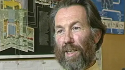The image is a close-up, indoor photograph, showing a single man positioned in front of wall-mounted educational display materials. Diagrams and printed charts are on the wall behind him. The man has medium-length hair brushed back from the forehead and a full beard and moustache extending across the cheeks and chin.
