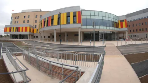 General view of Royal Stoke University Hospital. It is a large building with a semi-circular frontage with yellow, red and orange panels. There is a walkway in front of it.