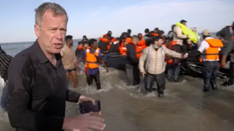 The BBC's Andrew Harding wearing a black shirt, standing in the sea, with a 'taxi-boat' full of migrants in the background. 