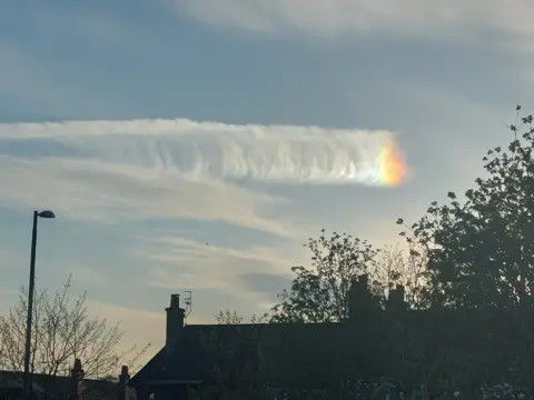 Ian Black A cloud with a rainbow reflection at the end. The silhouettes of houses and trees are in the foreground. 