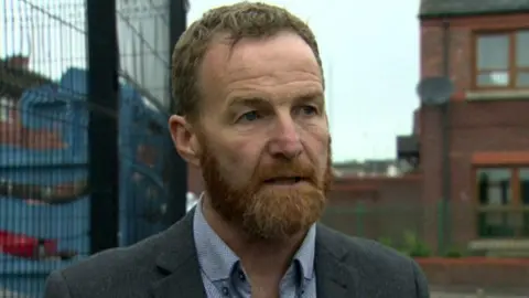 BBC A man in a grey jacket and blue shirt, with fair hair and a red beard stands in the street, in front of a black fence and red-brick house