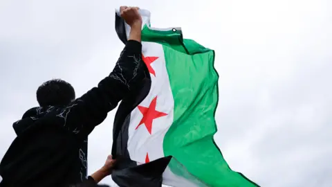 A man in an EPA sweatshirt, his back to the camera, waves in the air a Syrian opposition flag with green, black and white stripes with a red star. 