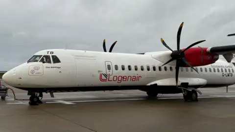 A white turboprop aircraft with a red tail and engine nacelles, parked on a wet airport apron under an overcast sky. The fuselage displays the word “Loganair” in large red letters with a stylized logo, and the text “Clan Donnachaidh” near the cockpit. The aircraft has two black propellers, and a ground power cable is connected to the nose gear area.