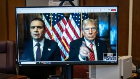 Getty Images Donald Trump and his lawyer Todd Blanche (left) appear virtually on a computer desktop screen from Florida for his sentencing in his Manhattan hush money case. Trump has his finger pointing at the camera, with US flags positioned behind him and Blanche.