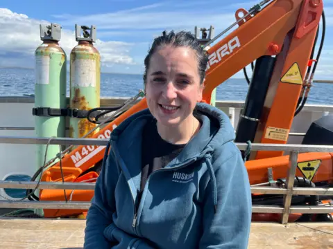 A woman with a blue jumper on, half-zipped up. She has dark hair which is tied back and is sitting in front of an orange crane, sea and cannisters.