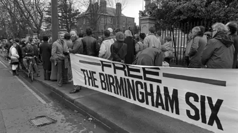 Getty Images Black and white image of a group of people standing on a pavement in front of a large banner that says 'Free the Birmingham Six', with houses, a metal fence and trees behind them.