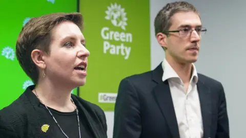 PA Media Carla Dener in a black jacket and t-shirt and Adrian Ramsau wearing a dark jecket and open necked white shirt stand on a stage with Green Party branding behind them
