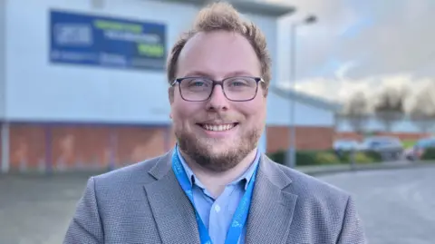 A man with fair hair and beard, wearing a grey blazer and blue lanyard, standing in front of a white cladded building with a brick base