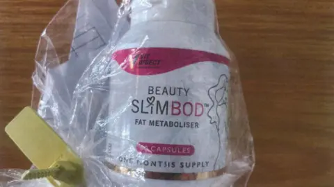 A clear plastic package laid on a wooden table with the a tub of the fake Slimbod pills inside. The packaging reads 'Beauty Slimbod Fat Metaboliser' - one month's supply. It is white and pink