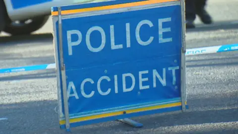 BBC Police accident sign, white writing on blue and yellow sign, with police tape and a police vehicle behind it