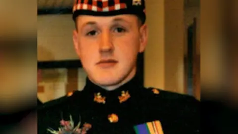 A young soldier in a Black Watch uniform and cap with red, white and black trim. He has medals and a flower on his uniform.