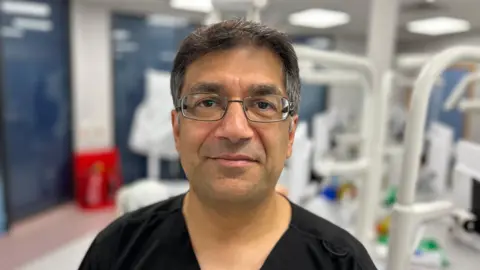 Dr Vishal Aggarwal, head and shoulders shot of an Asian man with glasses and short hair smiling at the camera in a dental room in a black top