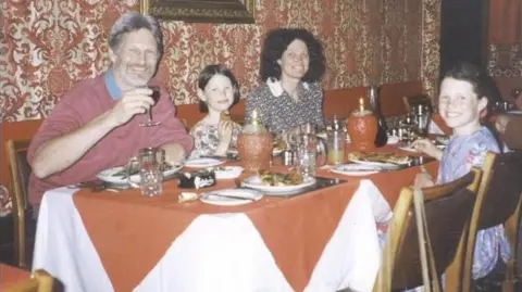 PA Media A man and woman sit either side of a young girl at a restaurant table. Another slightly older girl sits opposite them. They are all smiling. The man is holding a glass of what appears to be wine.