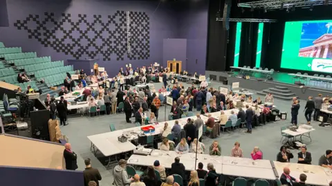 A widescreen shot, taken from a balcony, of the election count, with many people sat at tables and party members chatting. There is a stage on the right.