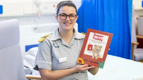 Anglia Ruskin University Ellie Falconer, who is wearing horn-rimmed glasses and has dark brown hair tied up, is dressed in a grey nursing uniform and is standing between a hospital bed and a chair, holding a copy of her book called Super Teddy. There is a blue curtain behind her.