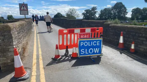 Warwickshire County Council The entrance to Bidford Bridge with orange cones blocking access to cars. A blue 'Police Slow' sign and an orange barrier are also in place. People are walking along the bridge, including a man walking a dog.