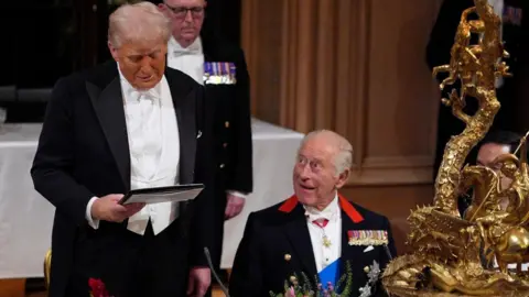 Getty Images President Donald Trump is stood at the table in a grand banquet hall wearing a tuxedo and reading something from papers held in his right hand. He appears to be smirking. King Charles is seated to his right, looking up at Trump.