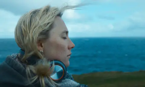 STUDIO CANAL A person with light hair stands outdoors near a large body of water, facing sideways with eyes closed.
The person is wearing layered clothing and has a pair of headphones resting around the neck.
Behind the person, the landscape shows a grassy shoreline and a wide, cloudy sky over the sea.